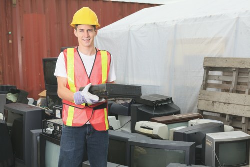 A young man wearing a yellow safety helmet, a high-visibility pink and yellow vest, and gloves stands outdoors in front of an array of discarded electronic waste, including old television sets and computer monitors with black or grey casings and glass screens. The background features a red wooden fence and a white tent, suggesting an outdoor waste collection or recycling site near Kilburn. The man is smiling and holding a black electronic device, possibly a keyboard or small electronic component, indicating involvement in rubbish disposal or recycling activities. The scene captures the practical and professional environment associated with waste management services offered by Business Waste Removal Kilburn in NW10, emphasizing proper handling of electronic rubbish in a tidy, organized manner.