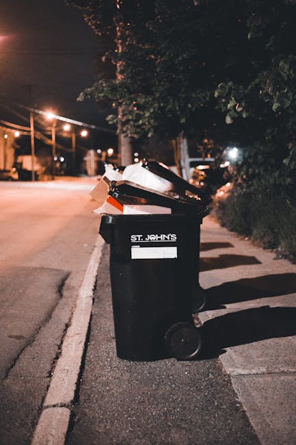 A black wheeled rubbish bin labeled with the name 'St. John's' stands on the edge of a quiet urban street at night, positioned on the pavement near the curb. The bin is partially open, revealing a mix