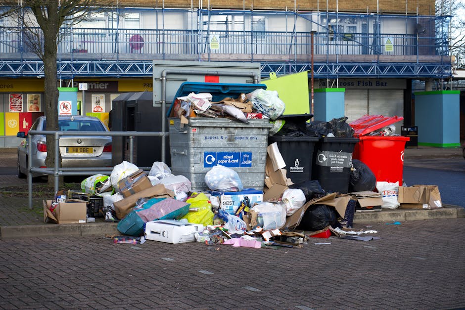 A busy street scene in front of a row of commercial premises, including a pharmacy and a seafood shop, with multiple people gathered around various items placed on the pavement. The foreground shows a